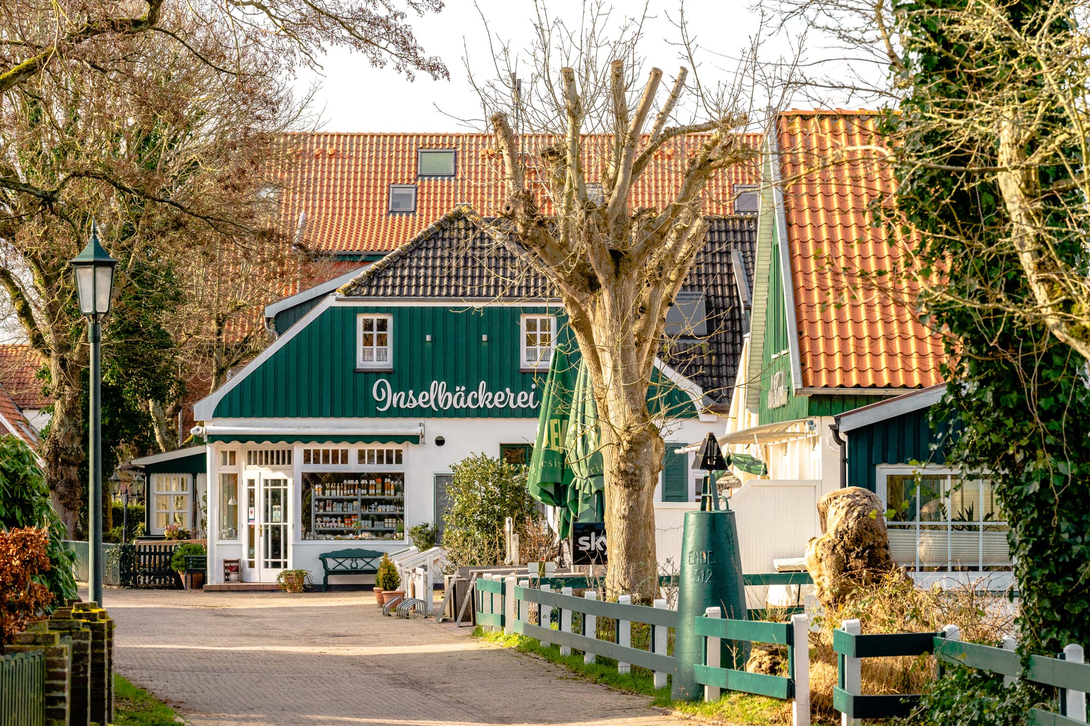 Historische Häuserzeile im Inseldorf Spiekeroog mit der Inselbäckerei und baumgesäumtem Weg Historische Häuserzeile im Inseldorf Spiekeroog mit der Inselbäckerei und baumgesäumtem Weg